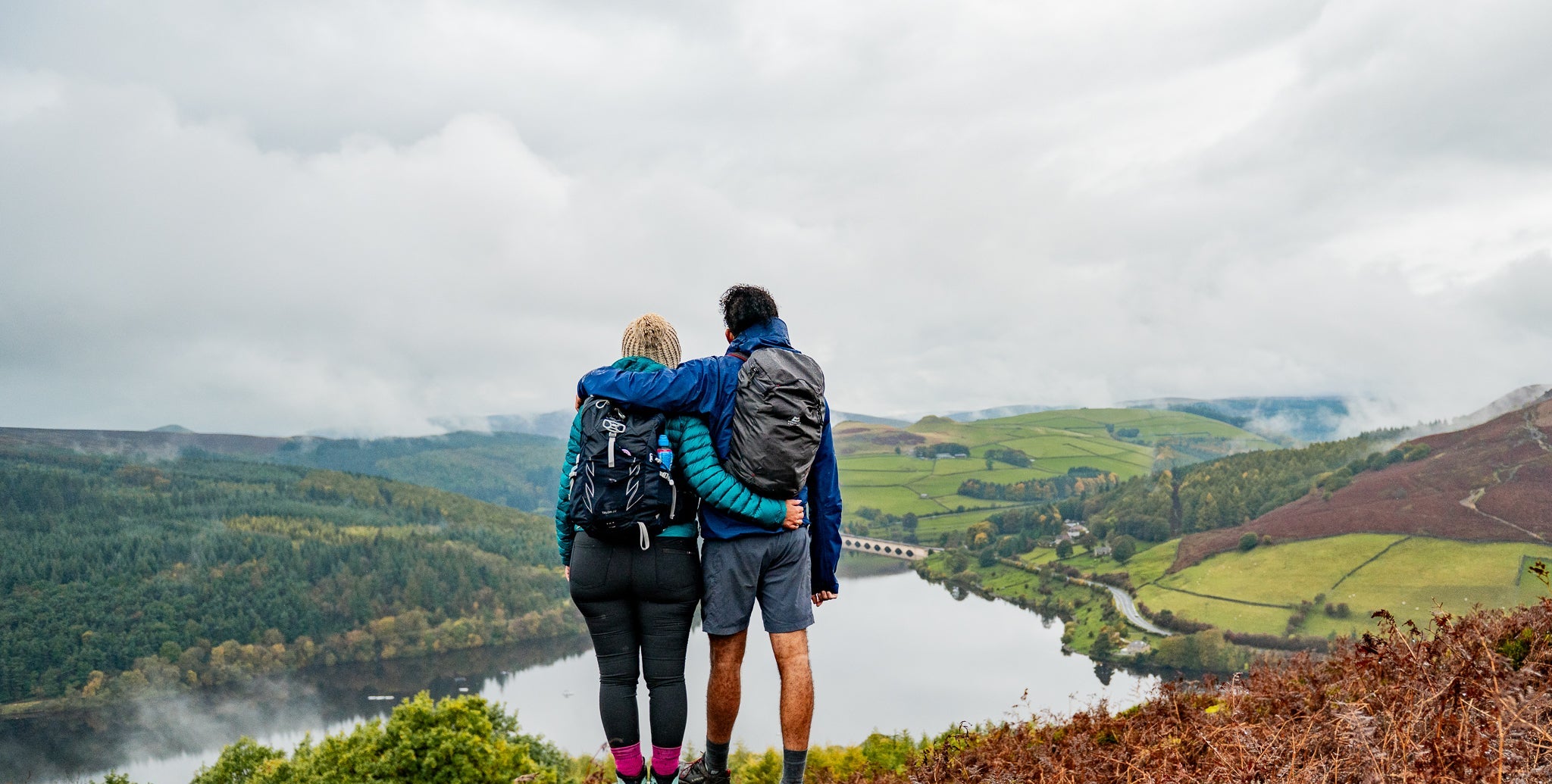 Two people standing on a rocky outcrop overlooking a scenic landscape with a lake and mountains.