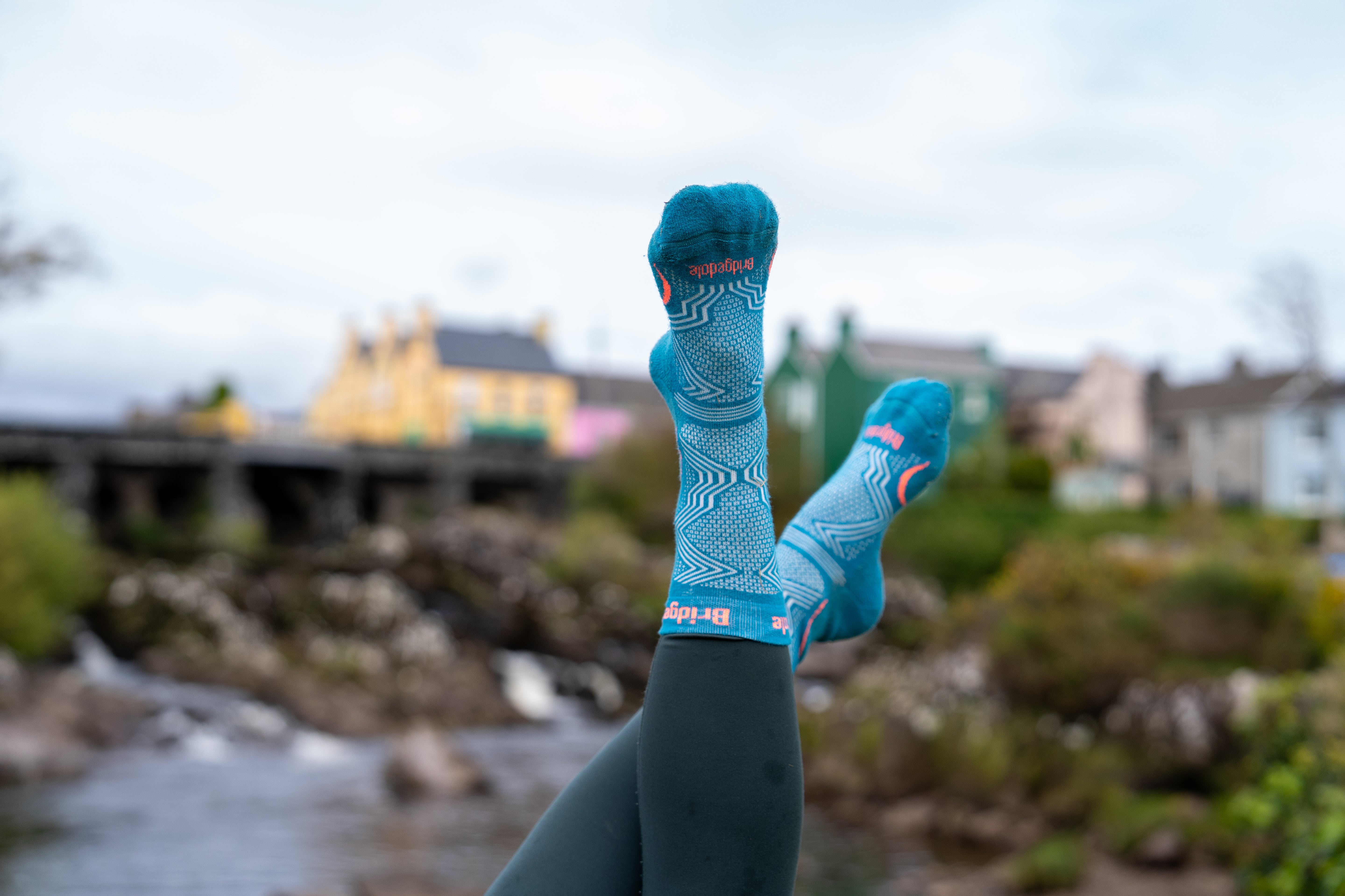 A person's legs raised in the air wearing bright blue and teal patterned athletic socks with dark leggings, set against a blurred outdoor background of a river and buildings.